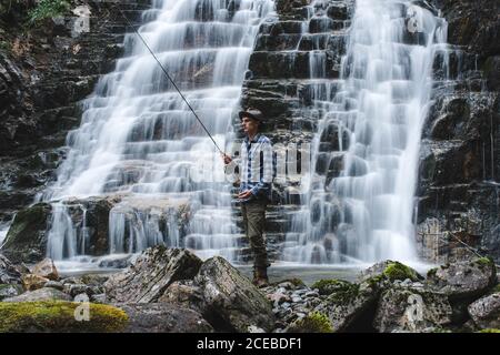 Fisherman dressed in fishing hat with blue shirt and gray marching trousers holding a spinning rod standing on rocks behind the slope of a waterfall Stock Photo