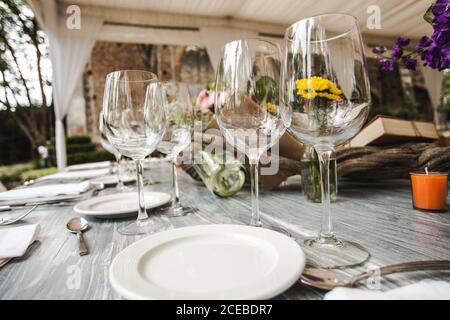 Fragile empty goblets standing near ceramic plates on beautifully decorated banquet table under tent Stock Photo