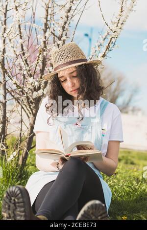 Trendy young Woman in hat sitting on green meadow with blooming tree reading book Stock Photo