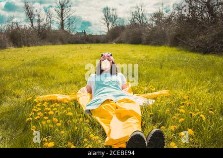 Young stylish Woman in raincoat and hat lying on green meadow with flowers and dreaming in sunlight Stock Photo