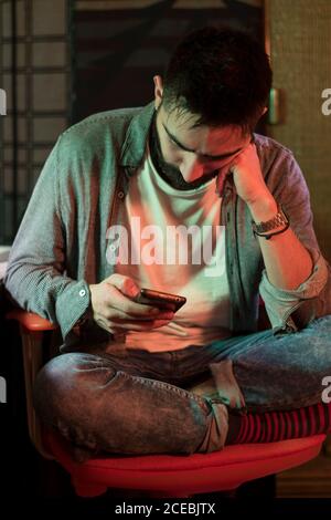 Thoughtful adult man sitting in chair and browsing smartphone indoors. Stock Photo