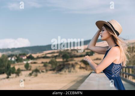 Side view of pretty young Woman holding hat and admiring view of magnificent countryside on windy day Stock Photo