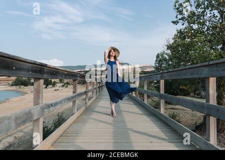 Attractive young female in elegant blue dress and hat walking along wooden path in wonderful countryside Stock Photo