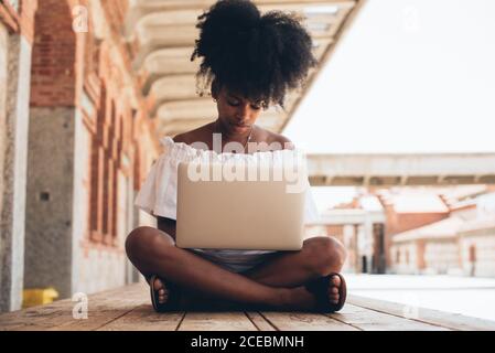 Pretty modern African-American Woman in white dress sitting on pavement with legs crossed and using laptop in sunlight Stock Photo