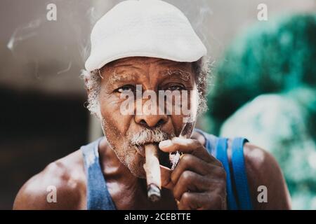 CUBA - September 14, 2017: Close-up view of cheerful senior Afro-Cuban male with large red flower on headdress holding cigar in mouth winking and looking at camera on blurred background Stock Photo