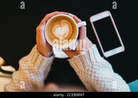 From above crop hands of lady in sweater holding mug of coffee near black table with mobile phone Stock Photo