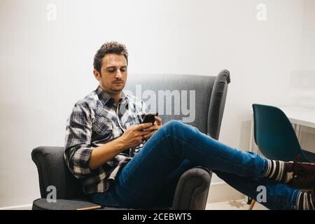 Young man using smartphone near laptop on armchair Stock Photo