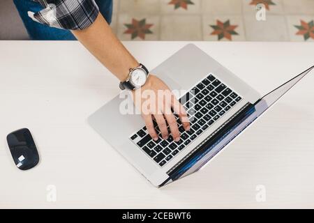 From above crop hand of male with watch using opened laptop near computer mouse on white table Stock Photo