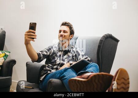 Smiling young man using smartphone near laptop on armchair Stock Photo