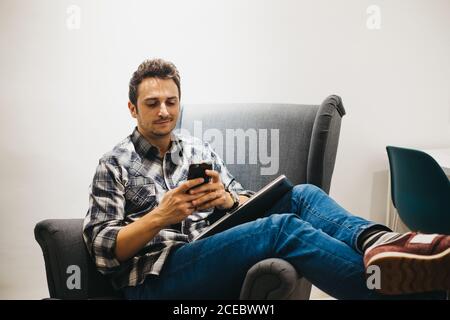 Smiling young man using smartphone near laptop on armchair Stock Photo