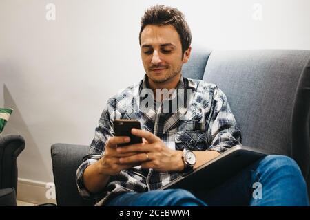 Pleased guy in casual wear with watch and mobile pone sitting on armchair with laptop on legs Stock Photo