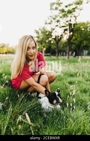 Attractive young blond lady in red dress sitting near French bulldog?with lead lying on green grass in park Stock Photo