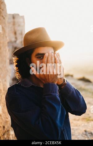 man in shirt and hat covering face with hands standing outdoors in bright sunshine against stone wall Stock Photo