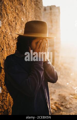 man in shirt and hat covering face with hands standing outdoors in bright sunshine against stone wall Stock Photo