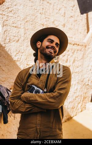 Casual bearded man in hat standing outdoors with photo camera on neck holding arms crossed and smiling away Stock Photo