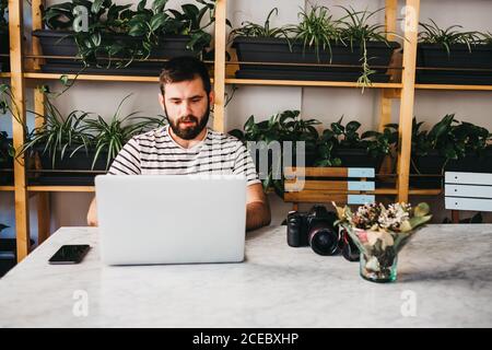 casual bearded man with photo camera and smartphone sitting at table while using laptop Stock Photo