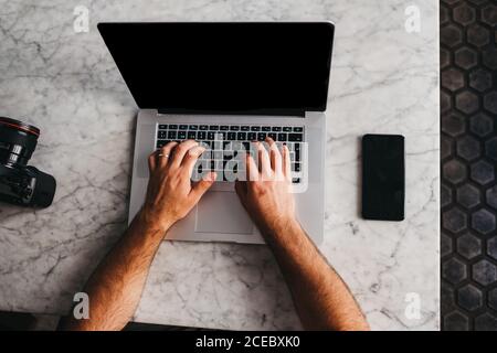 Crop shot from above of man using laptop sitting at marble table with phone near Stock Photo