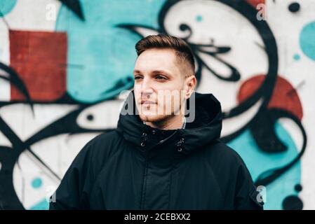 Thoughtful young man standing at graffiti wall with hands in pockets. Stock Photo