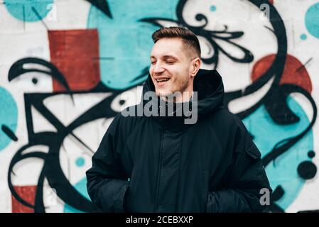 Thoughtful young man standing at graffiti wall with hands in pockets. Stock Photo