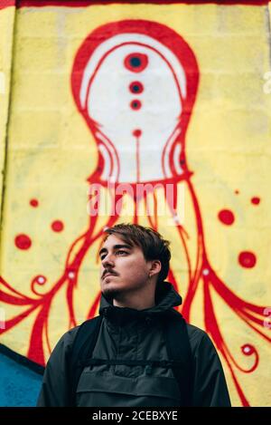 Thoughtful young man standing at graffiti wall with hands in pockets. Stock Photo