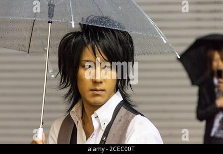 Japan - May, 10 2013: Stylish Asian guy with transparent umbrella looking away while standing on city street on rainy day Stock Photo