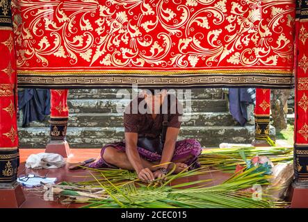 Bali, Indonesia - August, 15 2015: Adult ethnic male in traditional clothes braiding fresh palm leaves while sitting in ornamental tent near temple Stock Photo