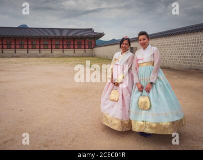 Seoul, South Korea - August, 08 2017: Two pretty Asian females in beautiful traditional apparels smiling and looking at camera while standing in courtyard on cloudy day Stock Photo