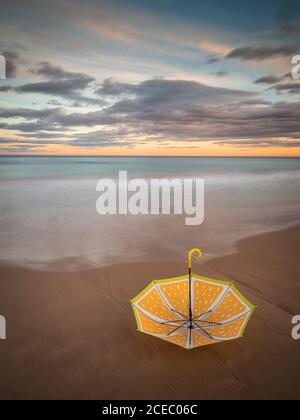 Cute yellow umbrella placed on sandy shore near waving sea during wonderful sunset in cloudy evening Stock Photo