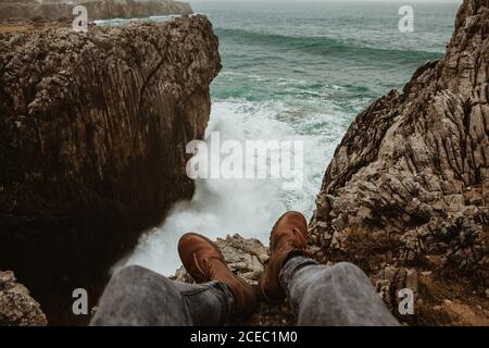 Crop legs of human sitting on top of stone near stormy sea in Bufones de Pria, Asturias, Spain Stock Photo
