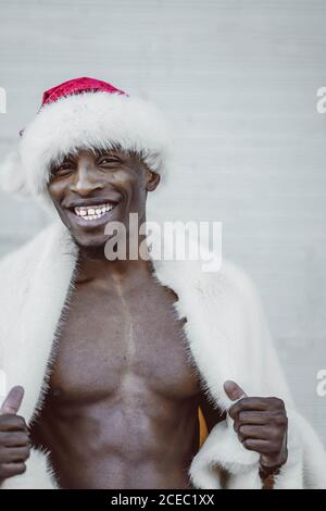 Handsome African American man in fur coat and Santa Claus hat smiling and looking at camera while standing near white wall Stock Photo