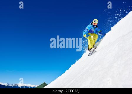 Male snowboarder riding snowboard fast down steep snowy mountain slope, jumping in air on copy space background of blue sky and white snow on sunny wi Stock Photo