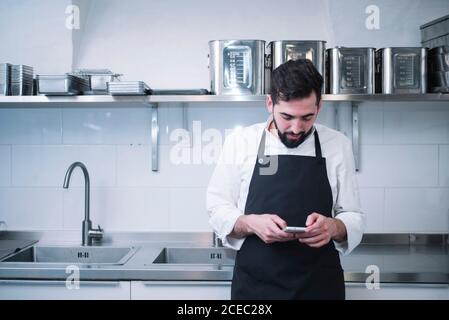 Side view of young man in cook uniform leaning on kitchen counter and using modern smartphone while standing in restaurant kitchen Stock Photo