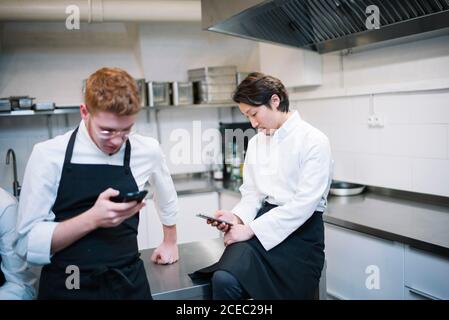 From below shot of two guys in cook uniform standing on restaurant kitchen and browsing smartphones during break Stock Photo