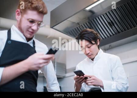 From below shot of two guys in cook uniform standing on restaurant kitchen and browsing smartphones during break Stock Photo