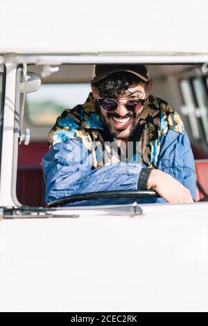 Attractive young man in stylish outfit leaning on steering wheel and looking at camera while sitting inside vintage van during trip in nature Stock Photo