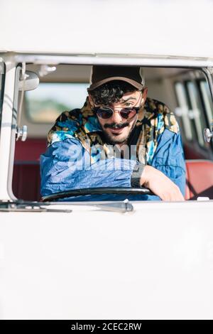 Attractive young man in stylish outfit leaning on steering wheel and looking at camera while sitting inside vintage van during trip in nature Stock Photo