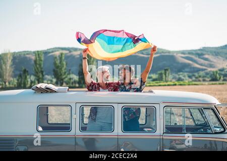 Two young men hugging and holding waving LGBT flag over heads while?standing inside retro van with opened roof in nature Stock Photo