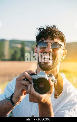 Handsome young guy in sunglasses cheerfully smiling and holding retro photo camera while standing on blurred background of amazing countryside Stock Photo