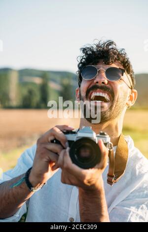 Handsome young guy in sunglasses cheerfully smiling and holding retro photo camera while standing on blurred background of amazing countryside Stock Photo