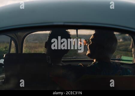 Back view of two unrecognizable young men hugging and admiring nature while sitting inside retro car Stock Photo