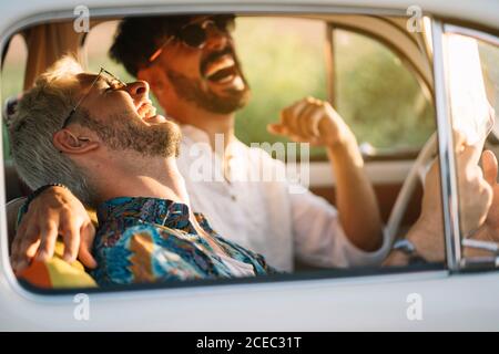 Two young guys laughing and holding map while sitting inside retro car during nice travel Stock Photo