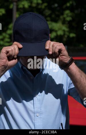 anonymous man in blue shirt wearing cap covering face while standing outside in sunlight