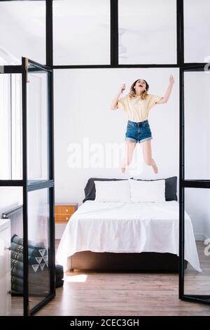 Young Woman in denim shorts jumping on bed happily holding phone and listening to music Stock Photo