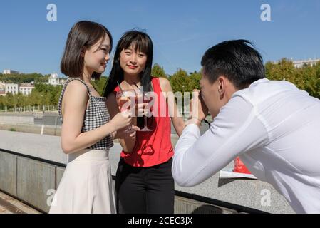 Young guy using camera to take pictures of two beautiful Asian ladies with glasses of fresh beverages while standing on city street on sunny day Stock Photo