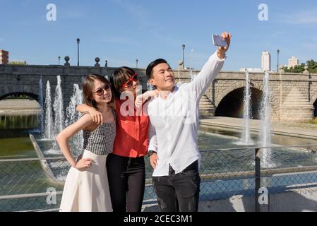 Cheerful Asian man and women in stylish outfits using smartphone to take selfie while standing near wonderful fountain on sunny day in park Stock Photo