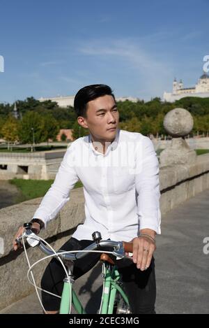 Handsome Asian guy looking away while riding bicycle along path in beautiful park Stock Photo