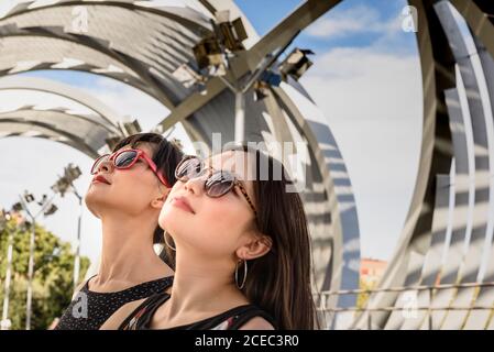 Two beautiful Asian ladies in stylish sunglasses looking up while standing inside modern structure on sunny day in park Stock Photo