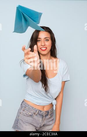 Young brunette smiling Woman throwing paper plane and looking at camera on blue background Stock Photo