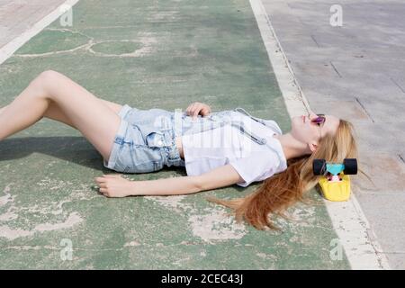 Side view of smiling girl in sunglasses and jeans overalls with white t-shirt lying on sidewalk with penny board under head Stock Photo