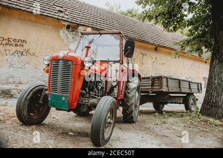 One old red tractor with trailer standing alone in front of building on the gravel street in shadow under the tree during summer sunny day in Serbia Stock Photo